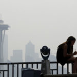 A girl works on a drawing next to an unused viewing scope as a smoky haze obscures the Space Needle and downtown Seattle behind, Tuesday, Aug. 14, 2018. Public-health officials are warning of unhealthy air across parts of the Pacific Northwest as smoke from wildfires move across the region. (AP Photo/Elaine Thompson)
