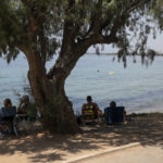 
              A man sunbathes as others sit under a pine tree at a beach of Kavouri suburb, southwest of Athens, on Friday, July 30, 2021. Greek authorities ordered additional fire patrols and infrastructure maintenance inspections Friday as the country grappled with a heat wave expected to last more than a week, with temperatures expected to reach 42 C (107.6 F). (AP Photo/Yorgos Karahalis)
            
