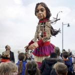 
              Actor Jude Law, center left, looks at Little Amal, a 3.5-meter-tall puppet of a nine-year-old Syrian girl, as it arrives at Folkestone Beach, Kent, England, Tuesday, Oct. 19, 2021 as part of the Handspring Puppet Company's 'The Walk'. The puppet started her journey in Turkey on 27 July and has travelled nearly 8,000 km across Greece, Italy, Germany, Switzerland, Belgium and France, symbolizing millions of displaced children. (Gareth Fuller/PA via AP)
            