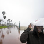 
              Michelle Jones covers her head with a bag as she watches large waves along Mission Beach Tuesday, Dec. 14, 2021, in San Diego. Rain is drenching Southern California as a powerful storm slides down the state, snarling traffic and raising the threat of mudslides in areas scarred by wildfires. (AP Photo/Gregory Bull)
            