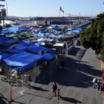 
              FILE - Migrants, below, walk outside a camp that blocks the entrance to a pedestrian crossing into the United States, above, Nov. 8, 2021, in Tijuana, Mexico. The encampment, now encircled in chain link fencing after an Oct. 28 operation by local police, is a temporary home for hundreds of migrants hoping to seek asylum in the United States. (AP Photo/Gregory Bull, File)
            