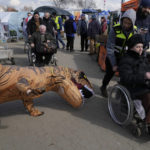 
              A volunteer dressed in a dinosaur suit jokes with a refugee in a wheelchair at the border crossing in Medyka, southeastern Poland, Monday, April 11, 2022. (AP Photo/Sergei Grits)
            