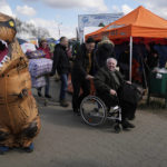 
              A refugee in a wheelchair smiles as a volunteer dressed in a dinosaur suit stands at the border crossing in Medyka, southeastern Poland, Monday, April 11, 2022. (AP Photo/Sergei Grits)
            