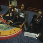 
              Rep. Tray McCurdy, D-Orlando, Rep. Angie Nixon, D-Jacksonville and Rep. Felicia Robinson, D-Miami Gardens sit on the Florida Seal in protest as debate stops on Senate Bill 2-C: Establishing the Congressional Districts of the State in the House of Representatives Thursday, April 21, 2022 at the Capitol in Tallahassee, Fla. The session was halted on the protest, but resumed later. (AP Photo/Phil Sears)
            