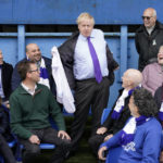 
              Britain's Prime Minister Boris Johnson removes his jacket as he is given a shirt with his name on, during a visit to Bury FC at their ground in Gigg Lane, Bury, Greater Manchester, England, Monday April 25, 2022. (Danny Lawson/Pool Photo via AP)
            