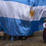 
              A child sits next to a giant national flag as a neighbor dressed as a gaucho waits to perform in a folkloric dance during the Independence Day celebrations in La Matanza, on the outskirts of Buenos Aires, Argentina, Wednesday, May 25, 2022. (AP Photo/Rodrigo Abd)
            