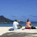 
              Brett Walsh, left, and Emma Yates, right, tourists from Australia, sit on Waikiki Beach in Honolulu, Monday, May 23, 2022. A COVID surge is under way that is starting to cause disruptions as schools wrap up for the year and Americans prepare for summer vacations. Case counts are as high as they've been since mid-February and those figures are likely a major undercount because of unreported home tests and asymptomatic infections. But the beaches beckoned and visitors have flocked to Hawaii, especially in recent months. (AP Photo/Caleb Jones)
            