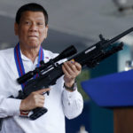 
              FILE - Philippine President Rodrigo Duterte jokes to photographers as he holds an Israeli-made Galil rifle at Camp Crame in suburban Quezon city northeast of Manila, Philippines on April 19, 2018. An International Criminal Court chief prosecutor, Karim Khan, said on June 24, 2022 that he has sought authorization from the court to resume an investigation into drug killings as a possible crime against humanity, from Nov. 1, 2011 when Duterte was still a Davao mayor to March 16, 2019. (AP Photo/Bullit Marquez, File)
            