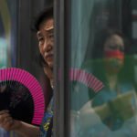 
              A woman fans herself at an advertisement boards on a bus stand reflecting a masked commuter as they wait for buses in Beijing, Tuesday, July 5, 2022. (AP Photo/Andy Wong)
            