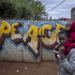 
              A boy carries a baby past a gate on which is written "Peace" in the Kibera neighborhood of Nairobi, Kenya Sunday, Aug. 14, 2022. Kenyans attended regular church services on Sunday, at which many pastors preached a message of patience and peace, as the country continues to wait for the results of Tuesday's presidential election. (AP Photo/Ben Curtis)
            