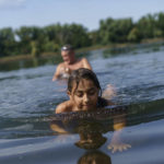 
              Anastasiia Aleksandrova, 12, plays in the water while swimming with her grandfather, Andreii, rear, at a lake in Sloviansk, Donetsk region, eastern Ukraine, Monday, Aug. 8, 2022. The mass displacement of Ukrainians, overwhelmingly women and children, has upended countless childhoods, not only for those having to start a new life after seeking safety elsewhere, but also for the thousands who have stayed behind. (AP Photo/David Goldman)
            