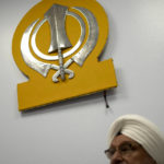 
              Paramjit Singh Bedi, co-chair of Guru Nanak Darbar of Long Island, a Sikh gurudwara, sits below the Khanda, the symbol of the Sikh faith, Wednesday, Aug. 24, 2022, in Hicksville, N.Y. An Afghan Sikh family of 13 has found refuge in the diaspora community on Long Island where the Sikh community is helping family members obtain work permits, housing, healthcare and find schools for the children. (AP Photo/John Minchillo)
            
