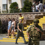 
              FILE - People walk past as Russian soldiers guard an office for Russian citizenship applications, in Melitopol, south Ukraine, July 14, 2022. This photo was taken during a trip organized by the Russian Ministry of Defense. (AP Photo, File)
            