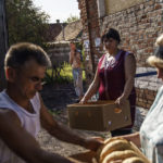 
              A resident waits in the background to buy bread as shopkeepers receive their daily delivery from Serhii Holoborodko, left, in Scherbynivka, Donetsk region, eastern Ukraine, Saturday, Aug. 20, 2022. By dawn, drivers arrive at the bakery to pick up fresh loaves of bread for delivery to towns and villages where the grocery stores are typically open only in the morning, when, on most days, there is a lull in Russian shelling. (AP Photo/David Goldman)
            