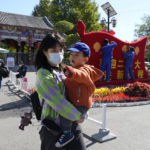 
              A woman wearing a mask carries a child past workers decorating a display celebrating the upcoming 20th Party Congress to be held in Beijing, Monday, Oct. 10, 2022. Chinese cities were imposing fresh lockdowns and travel restrictions after the number of new daily COVID-19 cases tripled during a weeklong holiday, ahead of the major Communist Party meeting in Beijing next week. (AP Photo/Ng Han Guan)
            