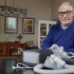 
              Jeffrey Reed, who experienced persistent sinus infections and two bouts of pneumonia while using a Philips CPAP machine, poses with the device at his home Thursday, Oct. 20, 2022, in Marysville, Ohio. The device is part of a massive global recall of breathing machines triggered by sound-dampening foam that can break down, releasing potentially harmful particles and chemicals. (AP Photo/Jay LaPrete)
            