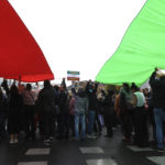 
              Protesters stand under an Iranian flag during a demonstration to show support for Iranian protesters standing up to their leadership over the death of a young woman in police custody, Sunday, Oct. 2, 2022 in Paris. Thousands of Iranians have taken to the streets over the last two weeks to protest the death of Mahsa Amini, a 22-year-old woman who had been detained by Iran's morality police in the capital of Tehran for allegedly not adhering to Iran's strict Islamic dress code. (AP Photo/Aurelien Morissard)
            