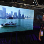 
              Samuel Seder, with NAVIER, stands next to video of the NAVIER 30 at the NAVIER booth during the CES tech show Friday, Jan. 6, 2023, in Las Vegas. The NAVIER 30 is the world's longest range electric hydrofoil boat. It flies 4.5 feet above the water. The NAVIER 30 is 10 times more efficient than a traditional gas boat. (AP Photo/Rick Bowmer)
            