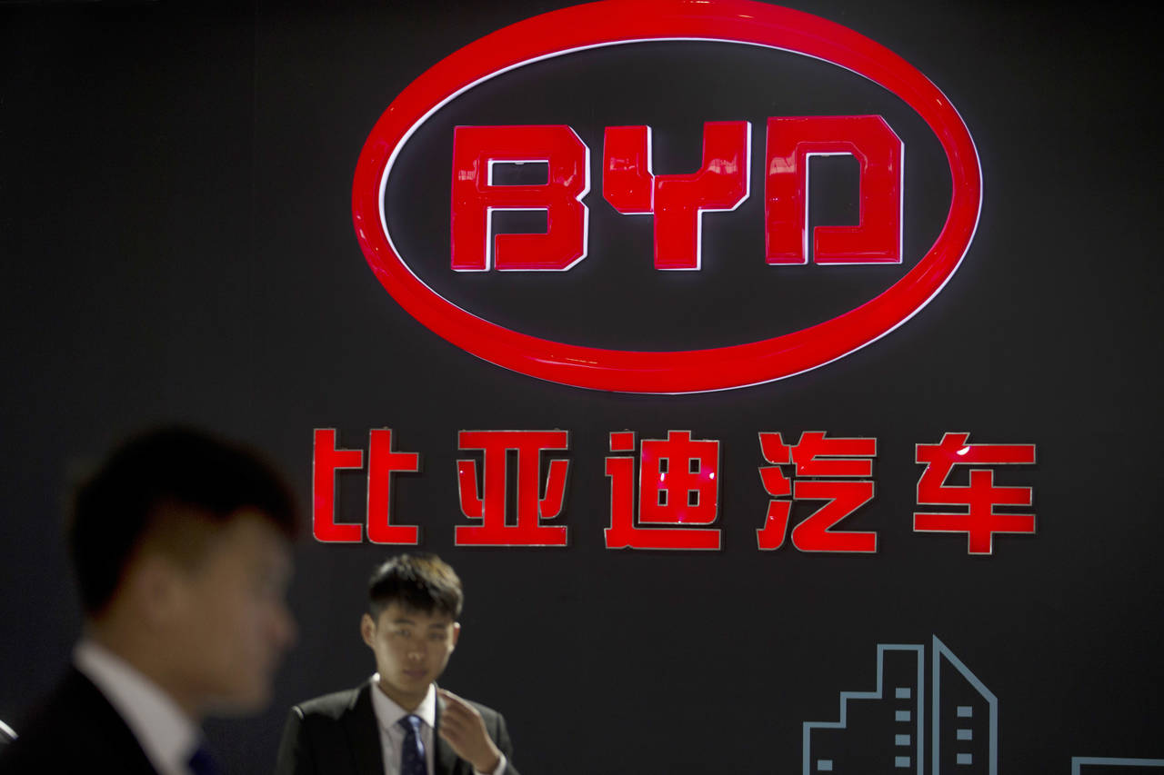 FILE - Staff members stand near a logo for the BYD auto company, as they wait for visitors at the B...