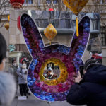 
              Women wearing face masks watch a toddler pose for a souvenir photo with a rabbit shaped floral decoration at a pedestrian shopping street at Qianmen on the first day of the Lunar New Year holiday in Beijing, Sunday, Jan. 22, 2023. People across China rang in the Lunar New Year on Sunday with large family gatherings and crowds visiting temples after the government lifted its strict "zero-COVID" policy, marking the biggest festive celebration since the pandemic began three years ago. (AP Photo/Andy Wong)
            