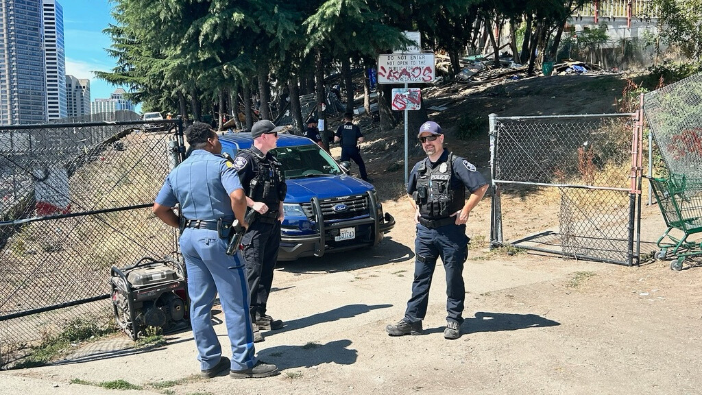 Image: Seattle Police Department officers stand at a homeless encampment just off of Interstate 5....