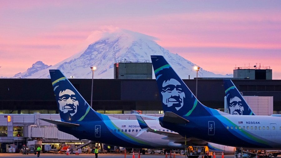 Image: Alaska Airlines planes are shown parked at gates with Mount Rainier in the background at sun...