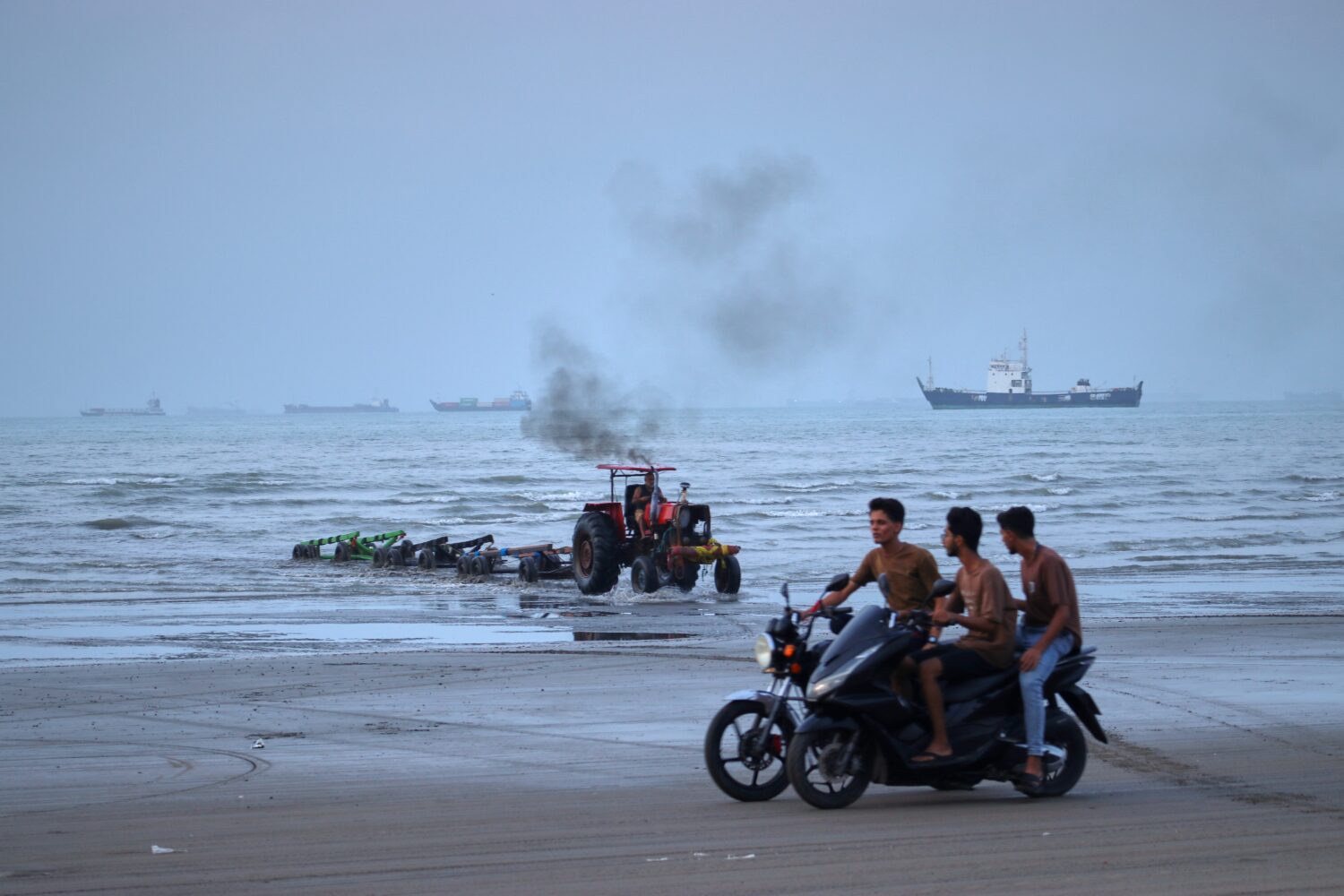 Young men ride motorcycles along the shore as a tractor pulls equipment from the water and commerci...
