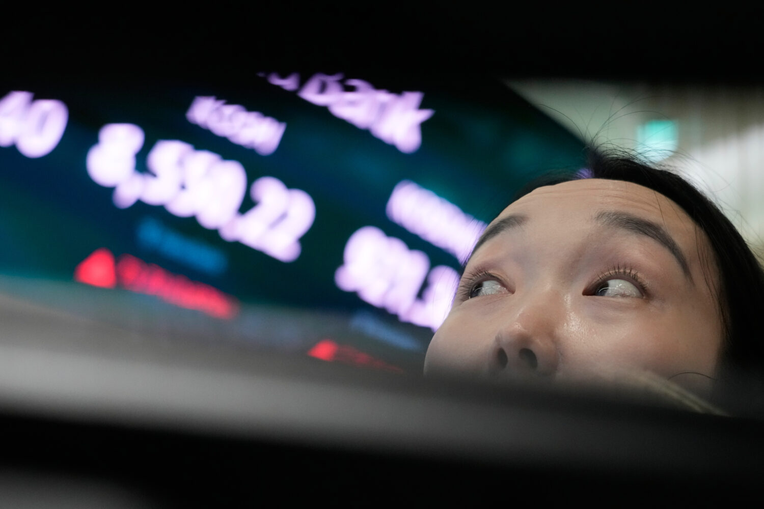 A currency trader watches monitors near a screen showing the Korea Composite Stock Price Index (KOS...