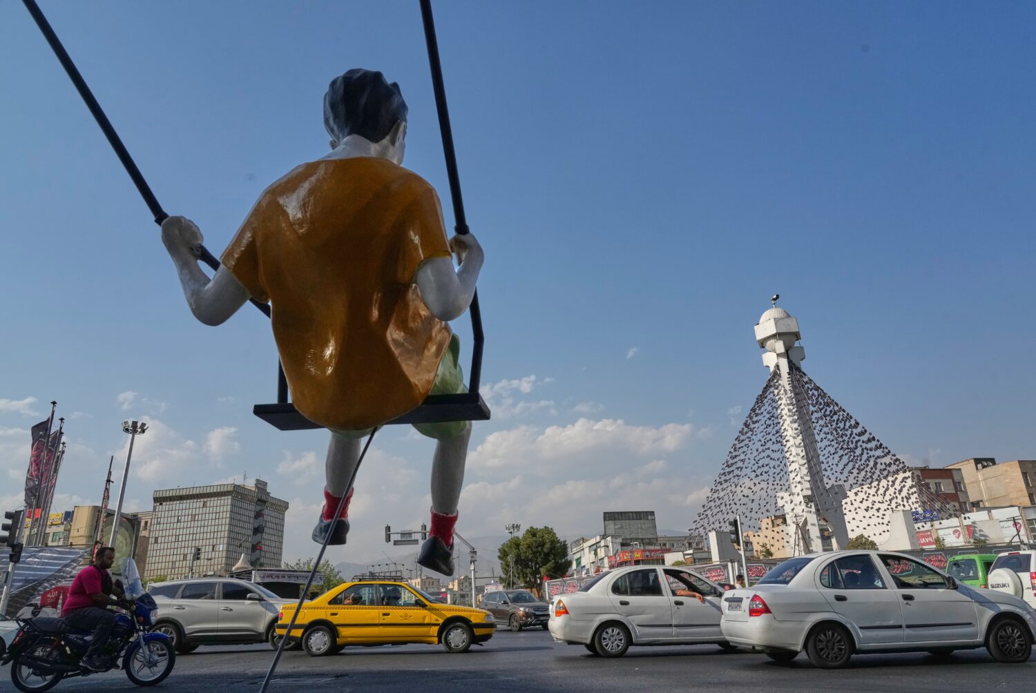 Vehicles drive past a statue of a swinging boy at a square in Tehran, Iran, Monday, Aug. 10, 2026. ...