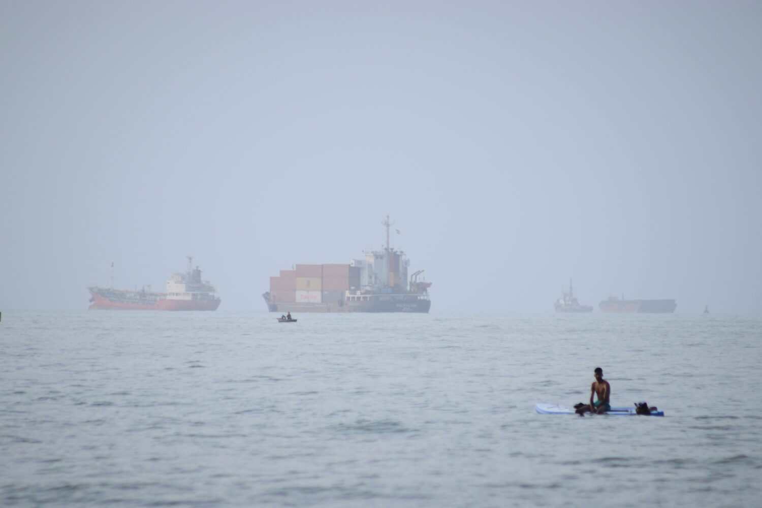 FILE - A man sits on a surfboard as a container ship and other commercial vessels appear anchored i...