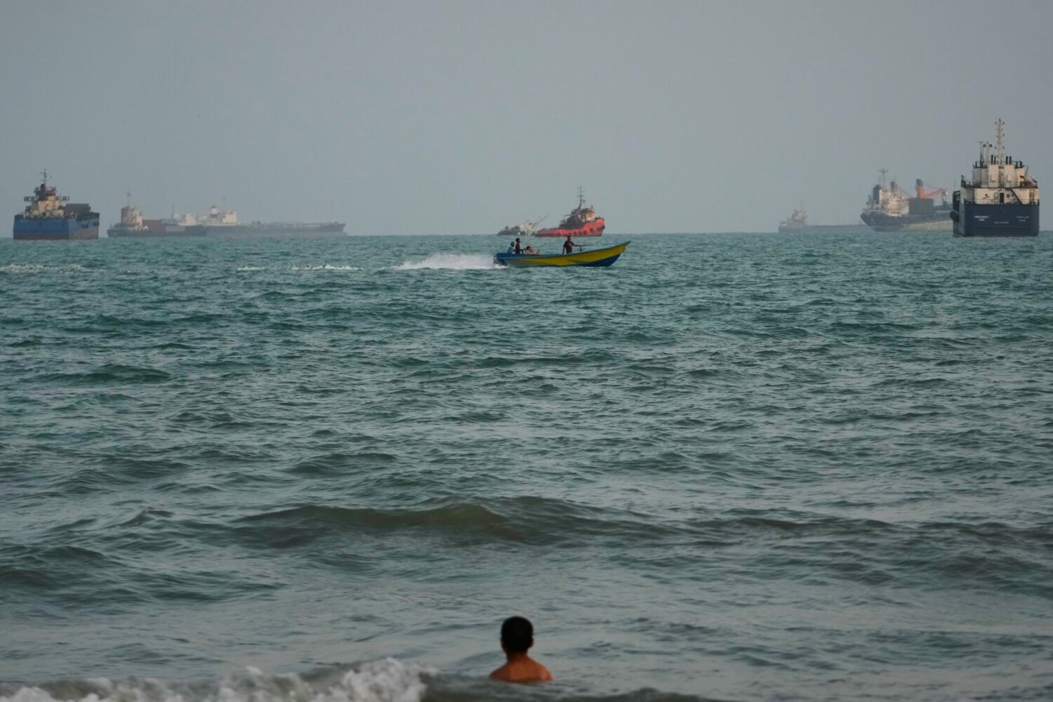 A swimmer remains near the shore as a motorboat passes commercial vessels anchored in the Strait of...