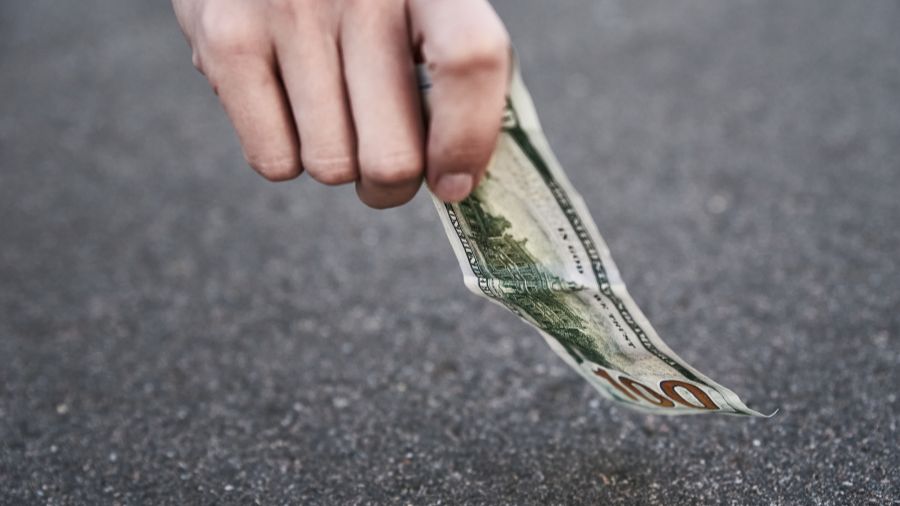 A hand holds a single one hundred US dollar bill over a grey asphalt surface. (Photo: Lazy_Bear via...