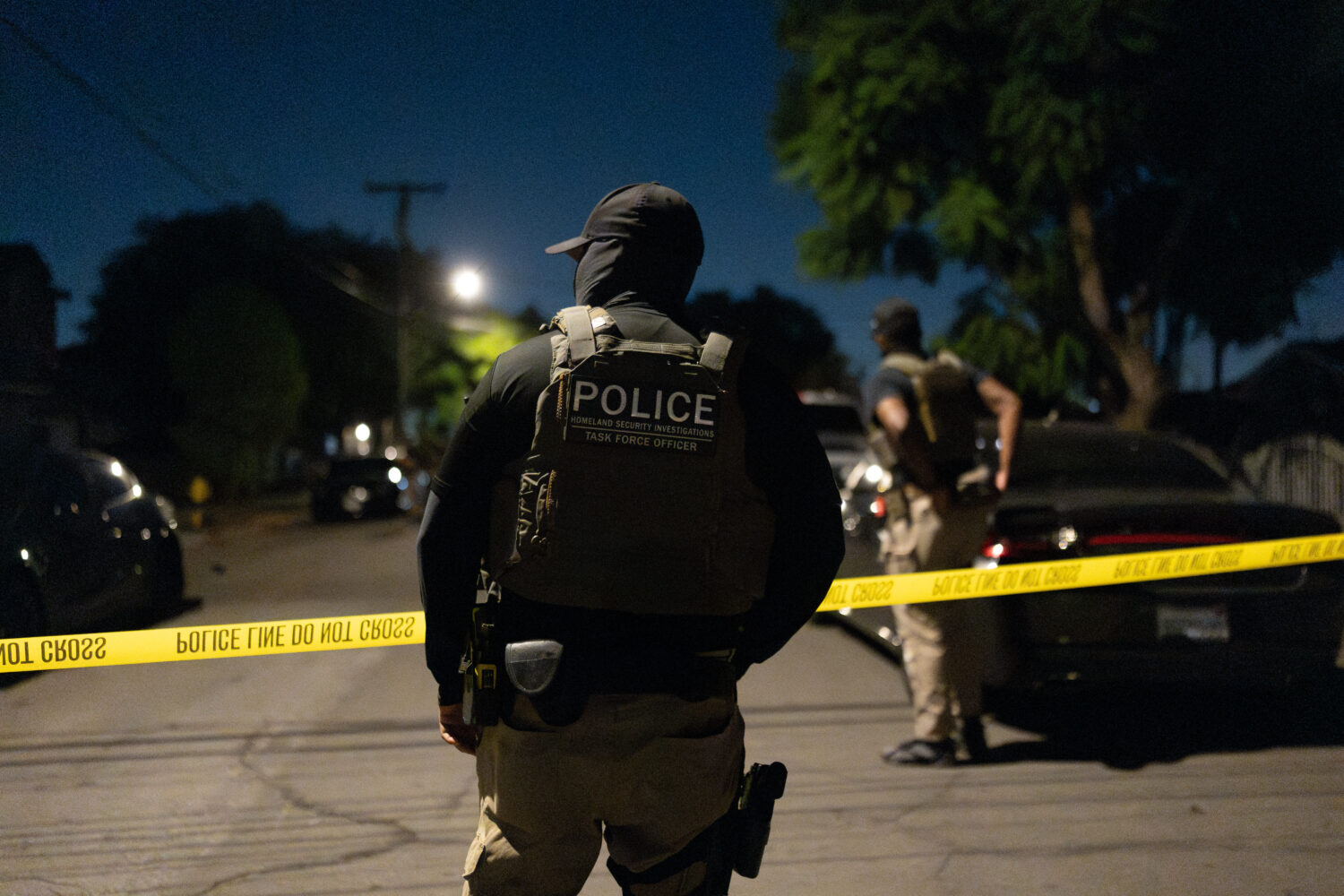 A Homeland Security Investigations task force officer stands near police tape marking a perimeter d...
