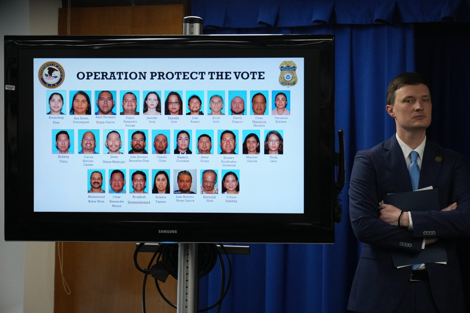 Acting U.S. Deputy Attorney General Trent McCotter stands next to a display showing photos of peopl...