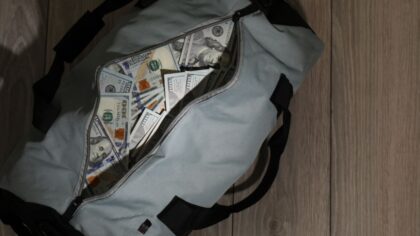 A partially unzipped grey duffel bag is overflowing with United States one hundred dollar bills. (P...