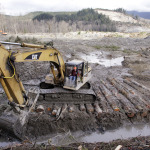 An excavator clears a drainage channel at the scene of a deadly mudslide, torn loose from the hillside at upper right, Wednesday, April 2, 2014, in Oso, Wash. The March 22 mudslide has killed at least 29 and another 13 are unaccounted for. (AP Photo/Elaine Thompson)