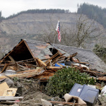 A flag sticks out of a hole in a demolished home near where a deadly mudslide from the hill behind that happened several days earlier ended, Tuesday, March 25, 2014, in Arlington, Wash. At least 14 people were killed in the 1-square-mile slide that hit in a rural area about 55 miles northeast of Seattle on Saturday. Several people also were critically injured, and homes were destroyed. (AP Photo/Elaine Thompson)